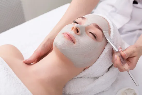 Woman receiving facial treatment with gray clay mask applied by beautician
